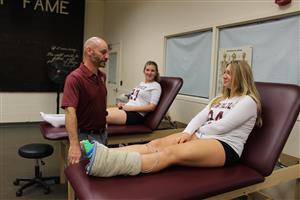 Athletic trainer works with injured athlete while other injured athlete looks on 