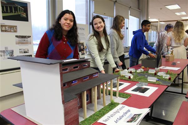 2 students behind table with projects smile while others look at items on table  